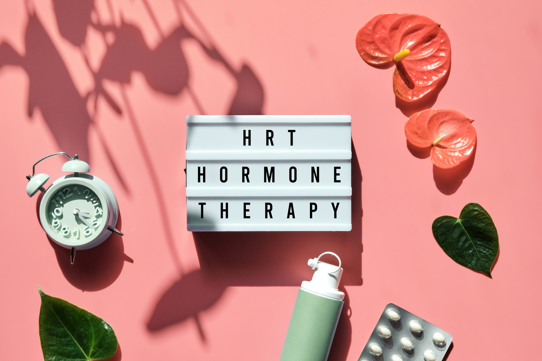 Text HRT Replacement Therapy on light box. Menopause, hormone therapy concept. Pink background with alarm clock, exotic leaves, pills, estrogene gel